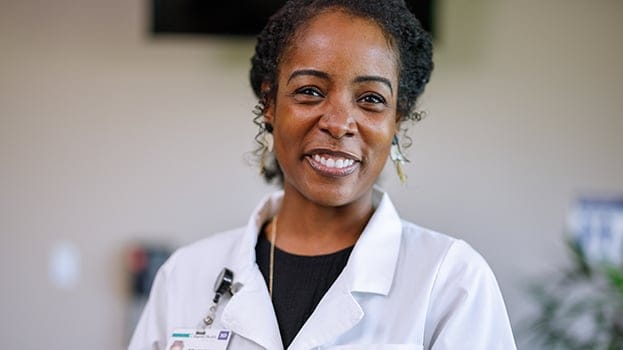 Smiling female doctor in a white coat standing in a hospital setting, representing the question “Who Is a Hospitalist.”