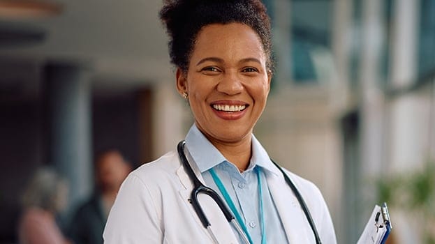 Smiling female doctor in a white coat with a stethoscope around her neck, holding a clipboard in a hospital setting.