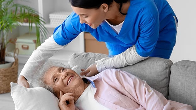 Smiling elderly woman lies on a couch while a nurse in blue scrubs leans over, gently adjusting her pillow.